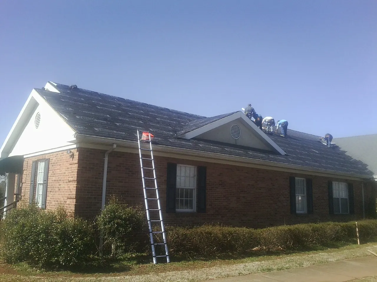 Crew tearing shingles off a brick ranch roof with a ladder set — Emergency Roof Repair work by our crew in Fort Harrison