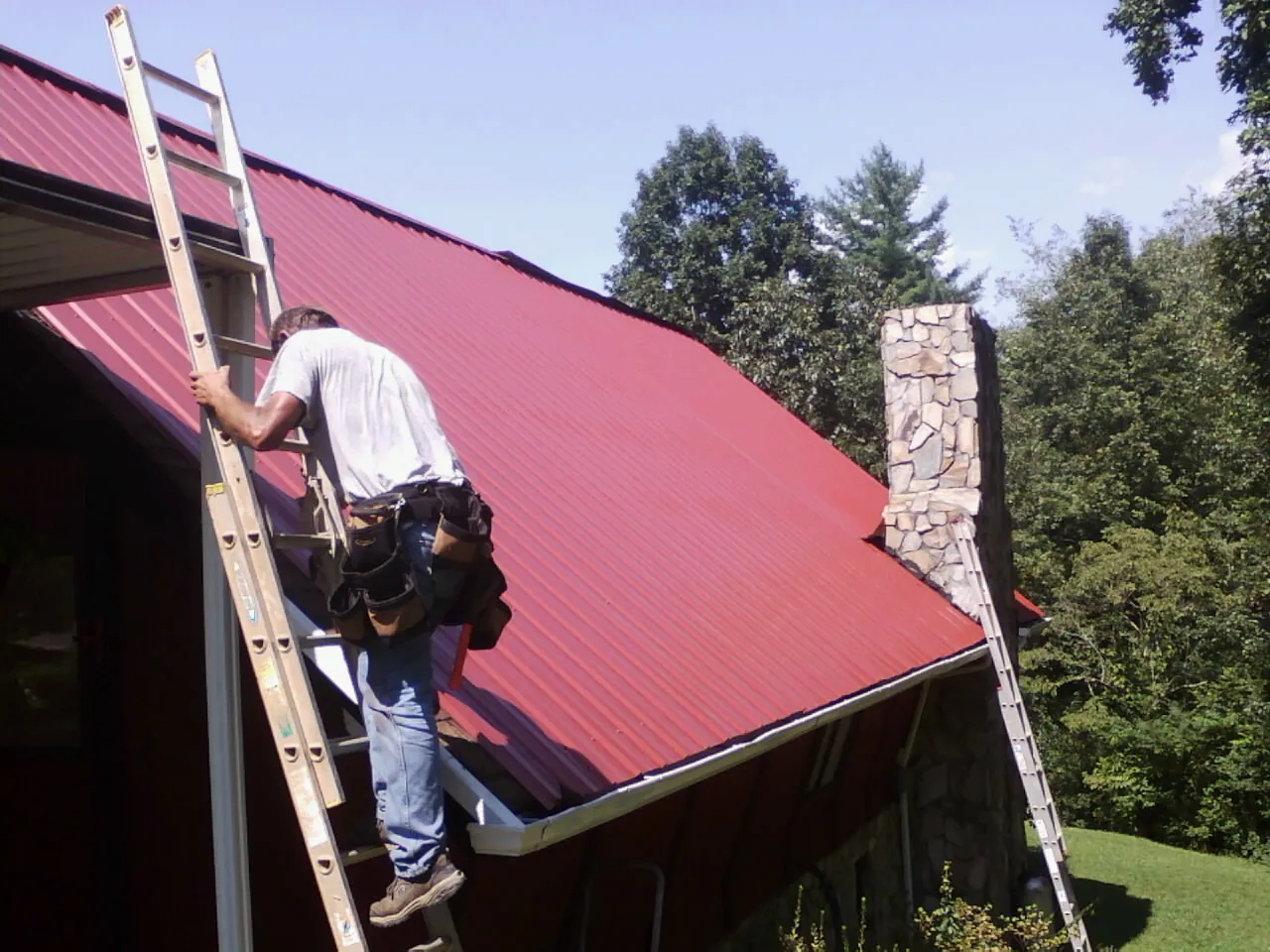 Roofer on a ladder beside a red metal roof and stone chimney — Soffit & Fascia Repair by Roofing Brothers in Fort Harrison